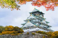A traditional Japanese castle with white walls and teal roofs sits atop a stone base, surrounded by vibrant foliage in shades of green, yellow, and red. The sky above is mostly cloudy with hints of blue peeking through.
