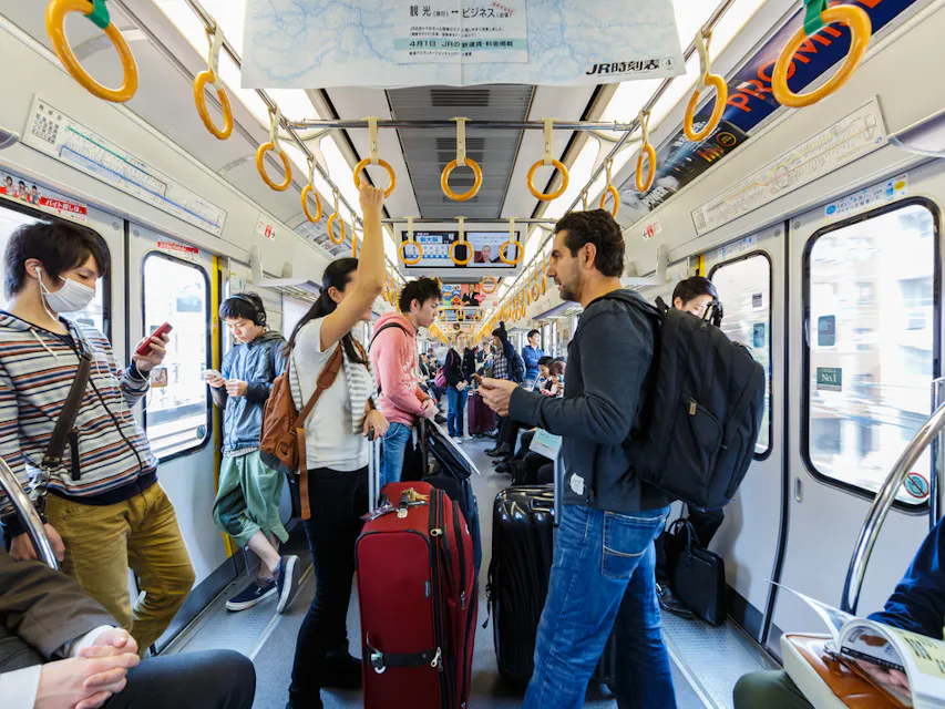 Subway Scene 乘客们在拥挤的火车车厢内站着和坐着,有些人抓着顶部的把手。行李放在地板上。人们在用手机和交谈。明亮的光线透过窗户,突出显示了外面繁忙的城市景象。