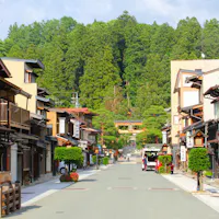 Takayama A serene street in a traditional Japanese town features wooden buildings with paper lanterns and potted plants along the sides. In the distance, a torii gate stands at the edge of a forest, framed by lush green trees under a partly cloudy sky.