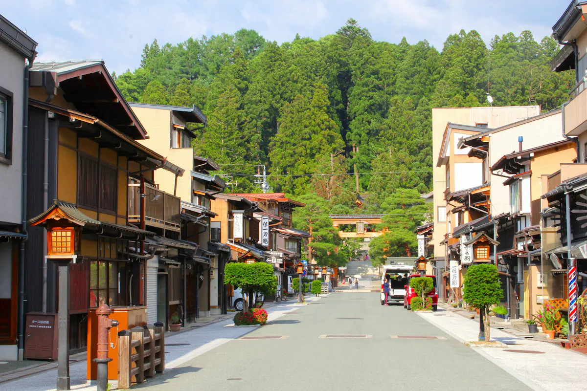Takayama A serene street in a traditional Japanese town features wooden buildings with paper lanterns and potted plants along the sides. In the distance, a torii gate stands at the edge of a forest, framed by lush green trees under a partly cloudy sky.