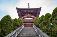 A traditional Japanese pagoda peeks between symmetrically aligned, manicured trees. The stone staircase leads up to the pagoda, showcasing its vibrant red wooden structure and intricate roofing details against a partly cloudy sky.