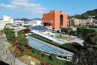 A modern campus with glass-roofed walkways, green trees, and a large red-brick building; distant mountains and a blue sky with scattered clouds in the background.