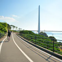 Cycling Routes in Japan A person rides a bicycle on a paved pathway surrounded by greenery and metal railings, with a tall cable-stayed bridge and blue sky in the background. The path runs alongside a body of water reflecting the sunlight.