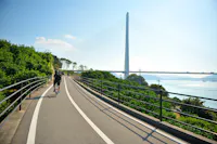 A person rides a bicycle on a paved pathway surrounded by greenery and metal railings, with a tall cable-stayed bridge and blue sky in the background. The path runs alongside a body of water reflecting the sunlight.