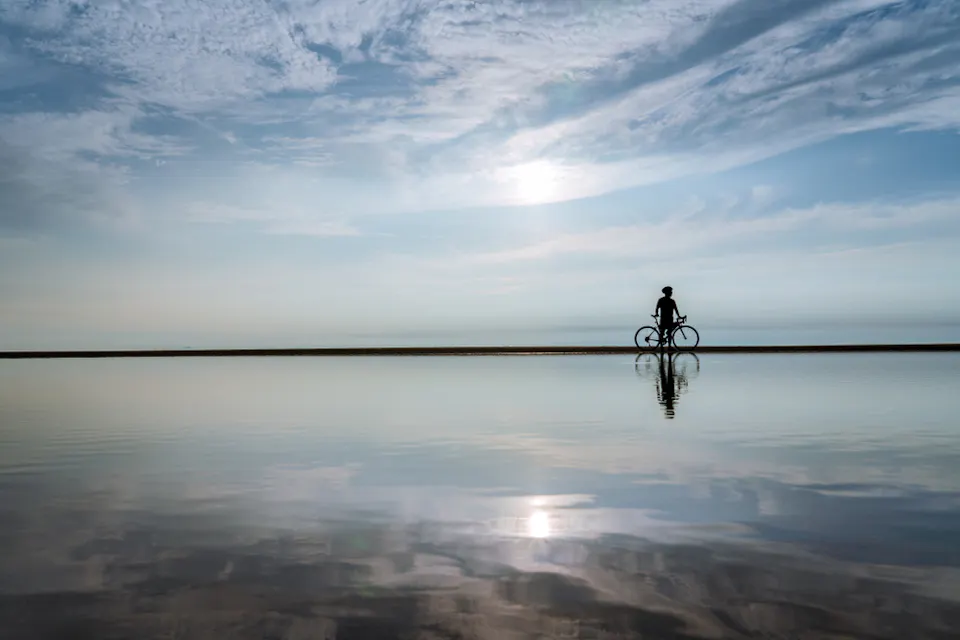Silhouetted figure riding a bicycle on a serene beach, with the sun low in the sky casting reflections on the calm water. The sky is filled with light, wispy clouds, creating a peaceful and tranquil scene.