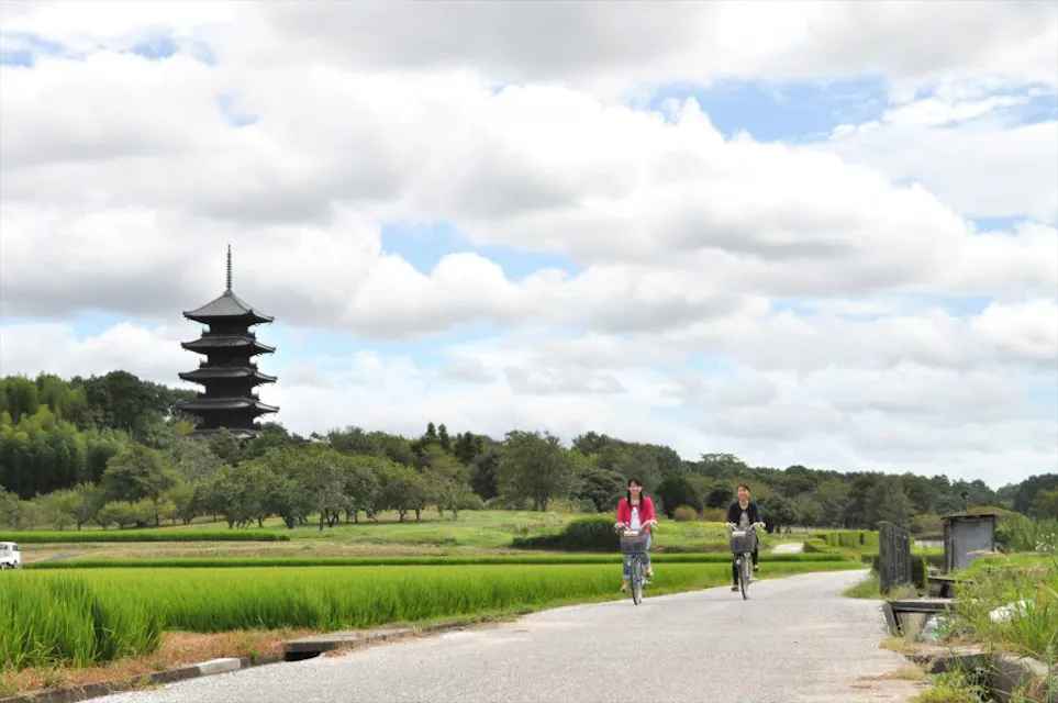 Two people ride bicycles on a paved path through lush green fields with scattered trees. A tall, multi-tiered traditional Japanese pagoda stands prominently in the background against a partly cloudy sky.
