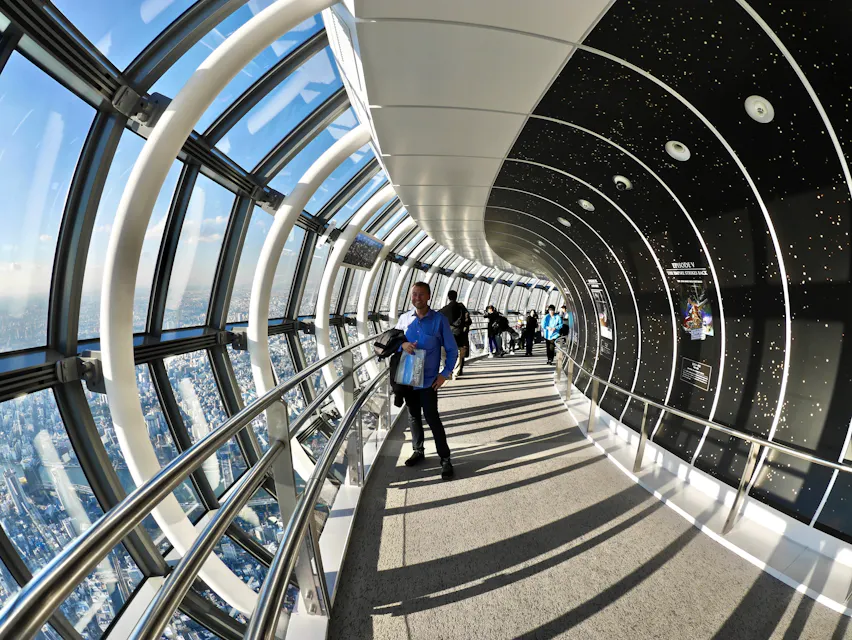 사람들이 곡선 유리 전망대 안을 걸으며 큰 창문을 통해 도시 풍경을 바라보고 있다. 내부는 벽에 별이 빛나는 하늘 패턴이 있고, 보행로는 금속 난간으로 둘러싸여 있다. 이 지역은 햇빛으로 밝게 비춰진다.