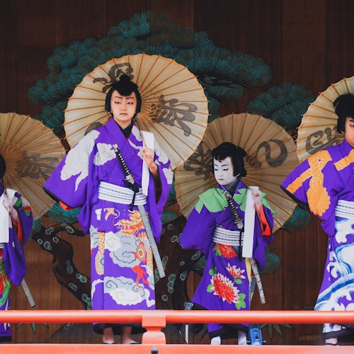 Kabuki A group of four performers in traditional Japanese attire stand on stage. They wear purple kimonos with elaborate designs and hold large decorated paper umbrellas. A painted backdrop of green trees is visible behind them.