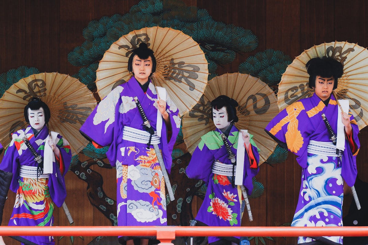 Kabuki A group of four performers in traditional Japanese attire stand on stage. They wear purple kimonos with elaborate designs and hold large decorated paper umbrellas. A painted backdrop of green trees is visible behind them.