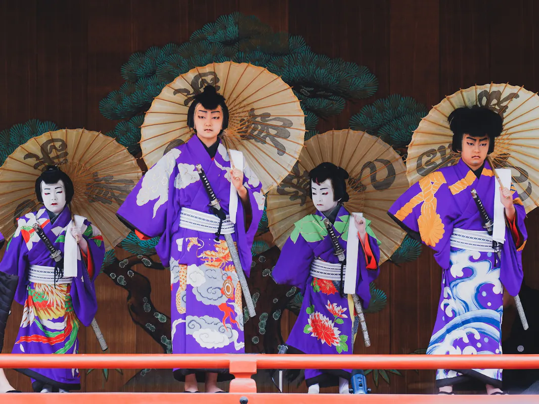 A group of four performers in traditional Japanese attire stand on stage. They wear purple kimonos with elaborate designs and hold large decorated paper umbrellas. A painted backdrop of green trees is visible behind them.