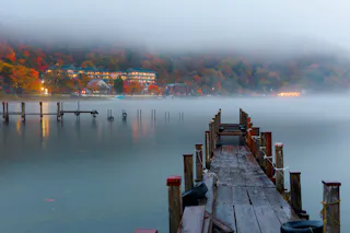 A wooden pier extends over a misty lake toward distant, tree-covered hills with autumn foliage. Buildings with warm lights are visible through the fog on the far shore, creating a tranquil, serene atmosphere.