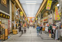A bustling indoor shopping street lined with various shops and stalls, selling clothes and other goods. Brightly colored banners and signs hang overhead. People can be seen walking, browsing, and shopping along the covered passageway.