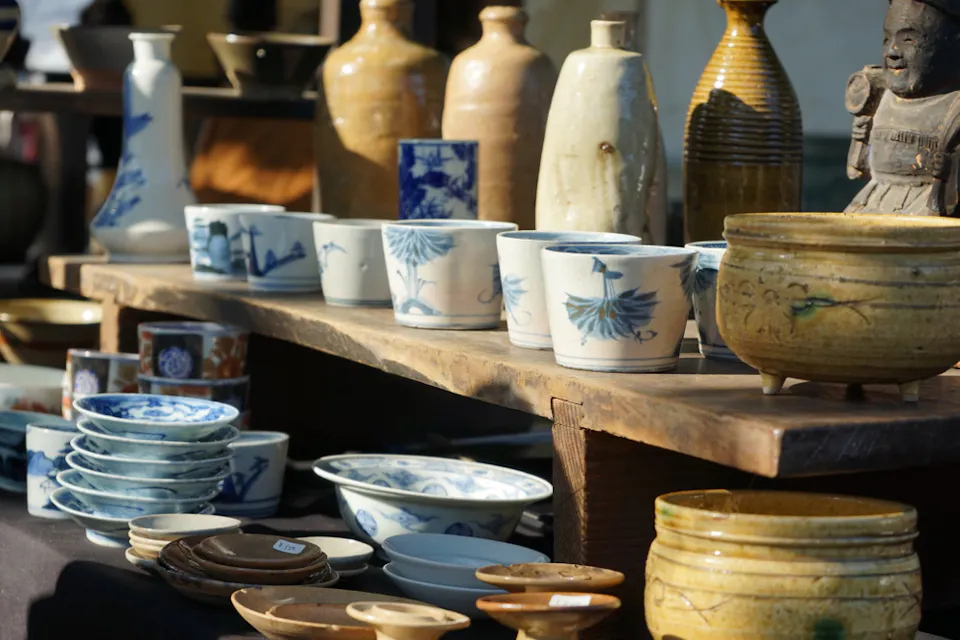 An array of ceramic items, including bowls, cups, plates, and vases, displayed on wooden shelves at an outdoor market. The ceramics feature various designs, with many showcasing blue and white patterns. A small statue is visible on the right side.