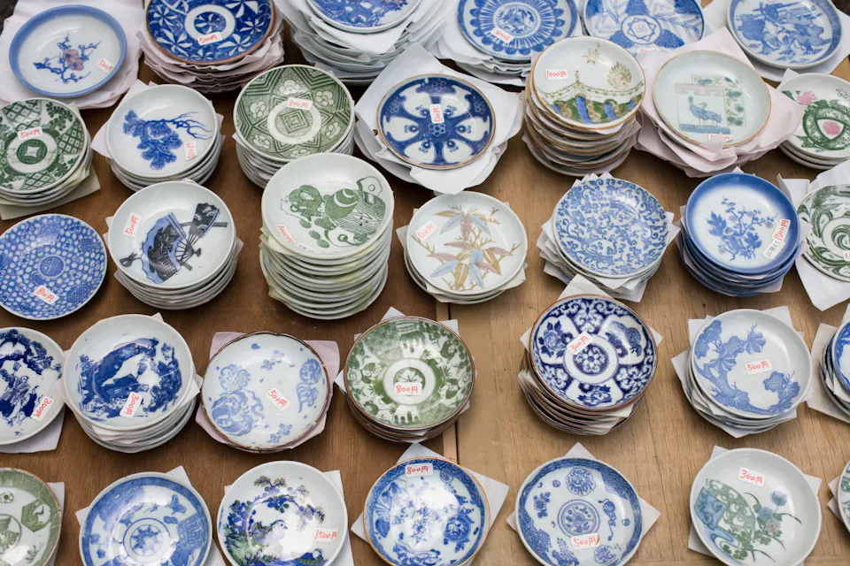 Stacks of various ceramic plates featuring intricate blue, green, and white patterns are displayed on a wooden surface. Many of the plates have small red price tags attached. The designs include floral, geometric, and animal motifs common in traditional pottery.