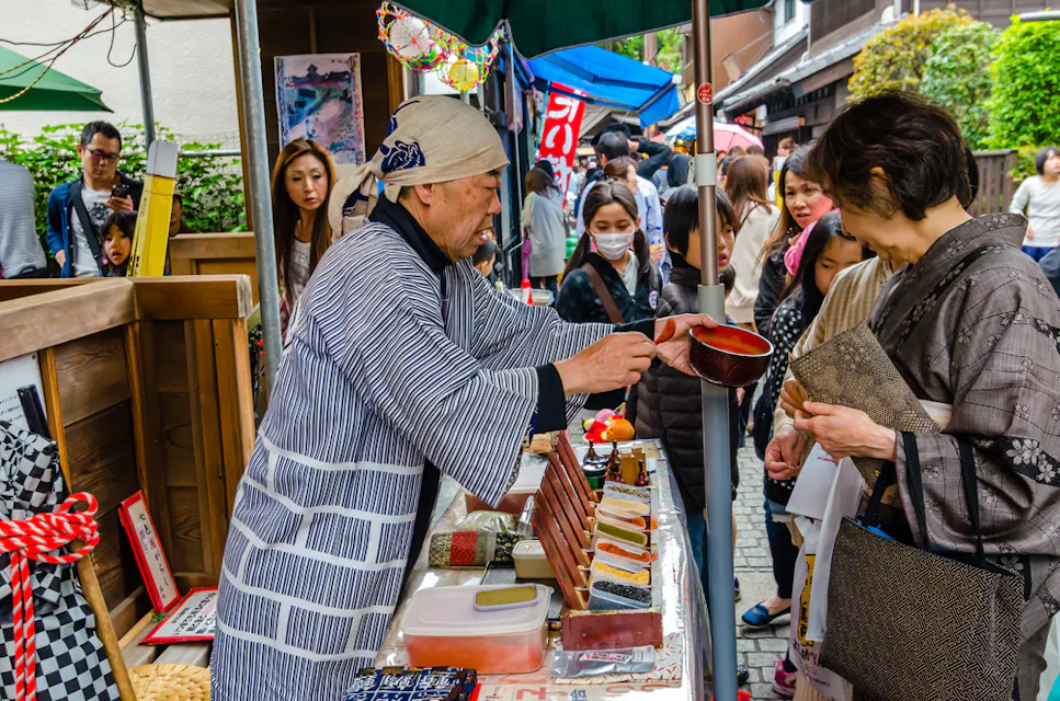 A vendor dressed in traditional attire serves food to a woman at an outdoor market. The market is bustling with people, colorful merchandise, and vibrant decorations. The vendor is standing behind a table filled with items, engaging with several onlookers.