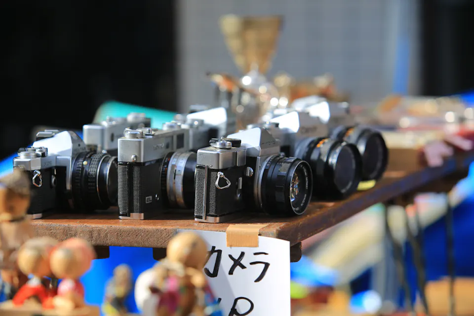 A collection of vintage cameras is displayed on a wooden table at an outdoor market. Below the cameras, miniature wooden dolls are scattered. The background features a blue sky, blurred to keep focus on the cameras. A handwritten sign with Japanese characters is visible.