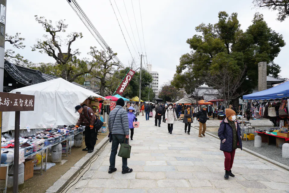 A busy outdoor market with various stalls lined up along a paved pathway. Shoppers browse items under white and blue canopies. Some stalls display colorful clothes, while others have accessories. Trees, buildings, and overcast sky are visible in the background.