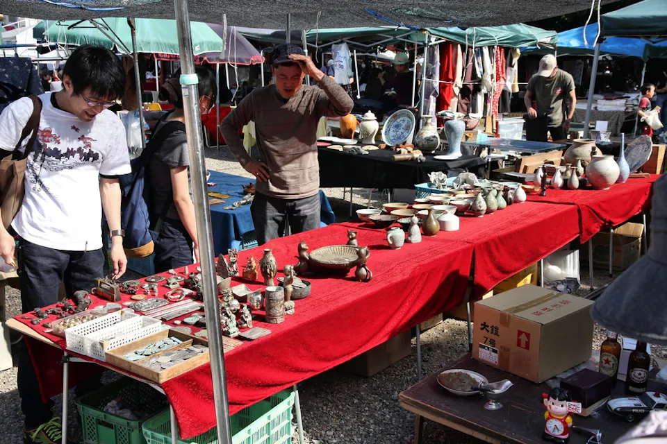 People browse various items displayed on tables covered with red cloth at an outdoor market. The tables are filled with pottery, figurines, and other trinkets. Vendors are present, and a variety of other market stalls can be seen in the background.