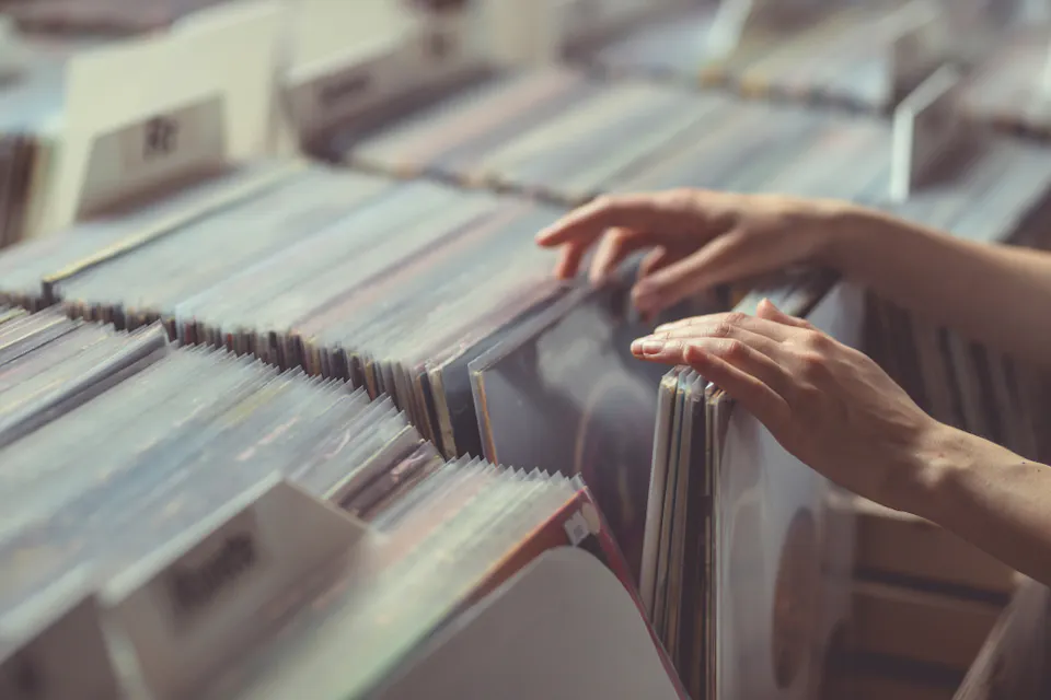 Two hands are seen browsing through a collection of vinyl records in a store setting. The image shows rows of vinyl records neatly organized in sleeves, with dividers. The overall atmosphere suggests a nostalgic or retro feel.