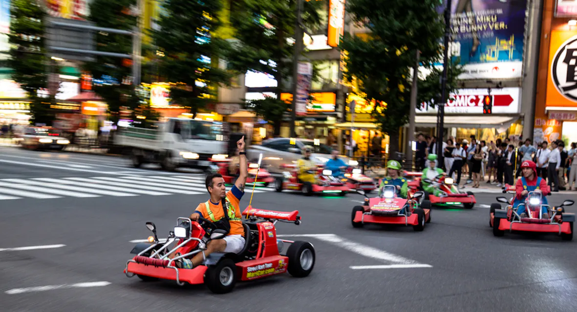 사람들이 도심의 밝은 불빛과 바쁜 횡단보도가 있는 거리에서 캐릭터 복장을 하고 고카트를 타고 있다. 이 장면은 실제 마리오 카트 레이스를 연상케 한다. 거리는 조명 광고로 둘러싸인 건물들로 둘러싸여 있다.