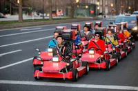 A group of people dressed in colorful outfits rides red go-karts on a city street. They are in two rows, smiling and enjoying their drive. The street has trees and buildings in the background, with evening lights adding a warm glow.