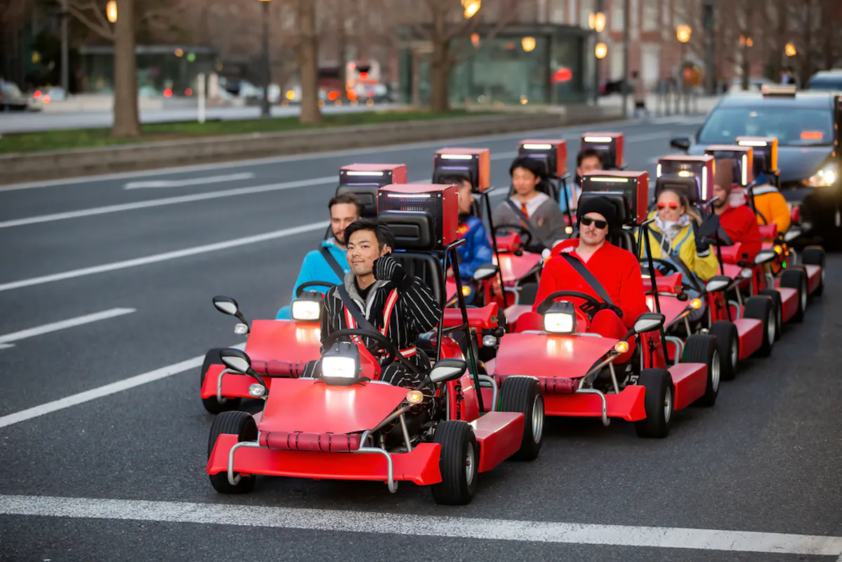 A group of people dressed in colorful outfits rides red go-karts on a city street. They are in two rows, smiling and enjoying their drive. The street has trees and buildings in the background, with evening lights adding a warm glow.