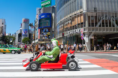 A person dressed in a green and white costume drives a red go-kart across a busy city street. The background features a large building with "TSUTAYA" signage, along with several pedestrians, vehicles, and billboards in an urban environment.