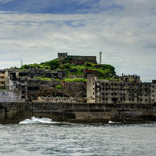 Hashima Island (Gunkanjima, Battleship Island) A coastal view of Hashima Island, also known as Battleship Island, featuring abandoned concrete buildings and a central green hill with a tall white tower. The sky is cloudy and there are waves crashing against the island's sea wall.