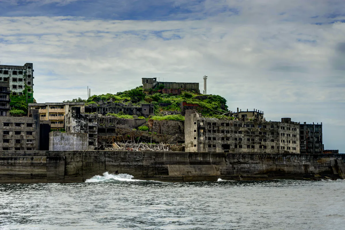 A coastal view of Hashima Island, also known as Battleship Island, featuring abandoned concrete buildings and a central green hill with a tall white tower. The sky is cloudy and there are waves crashing against the island's sea wall.