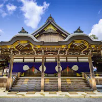 Ise Grand Shrine A traditional Shinto shrine with intricate wooden architecture stands under a clear blue sky. The structure features ornate, curved roofs with decorative elements, and steps leading to a platform adorned with white symbols on purple drapes. Trees surround the shrine.