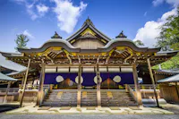 A traditional Shinto shrine with intricate wooden architecture stands under a clear blue sky. The structure features ornate, curved roofs with decorative elements, and steps leading to a platform adorned with white symbols on purple drapes. Trees surround the shrine.