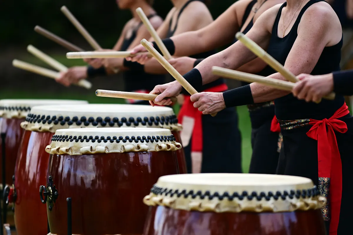 Sado's Taiko Traditions A group of people dressed in black tank tops and red sashes play traditional Japanese taiko drums. They are positioned in a row, striking the drums with thick wooden sticks, their arms raised mid-movement. The drums are large and feature decorative rope bindings.