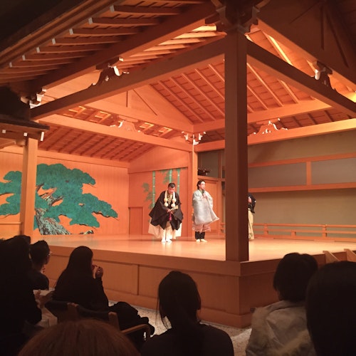 Noh Theatre A traditional Japanese Noh theater performance is taking place on a wooden stage with actors dressed in traditional attire. The backdrop features a green painting of a tree. An audience watches attentively from their seats.