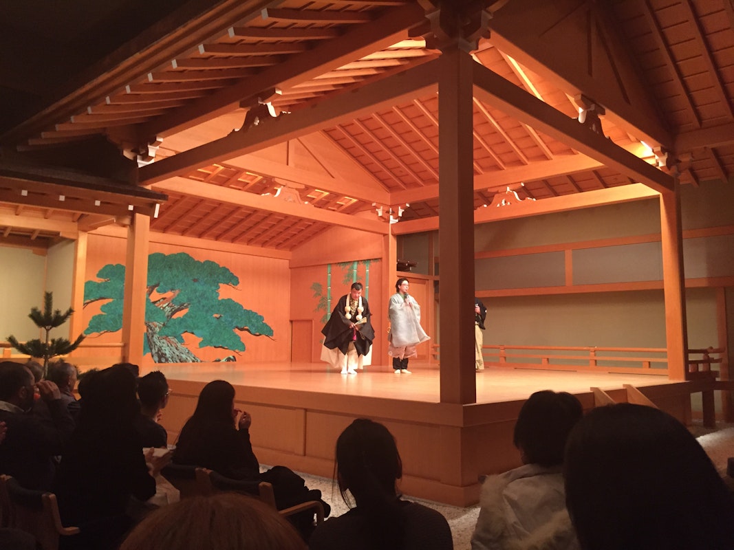 A traditional Japanese Noh theater performance is taking place on a wooden stage with actors dressed in traditional attire. The backdrop features a green painting of a tree. An audience watches attentively from their seats.