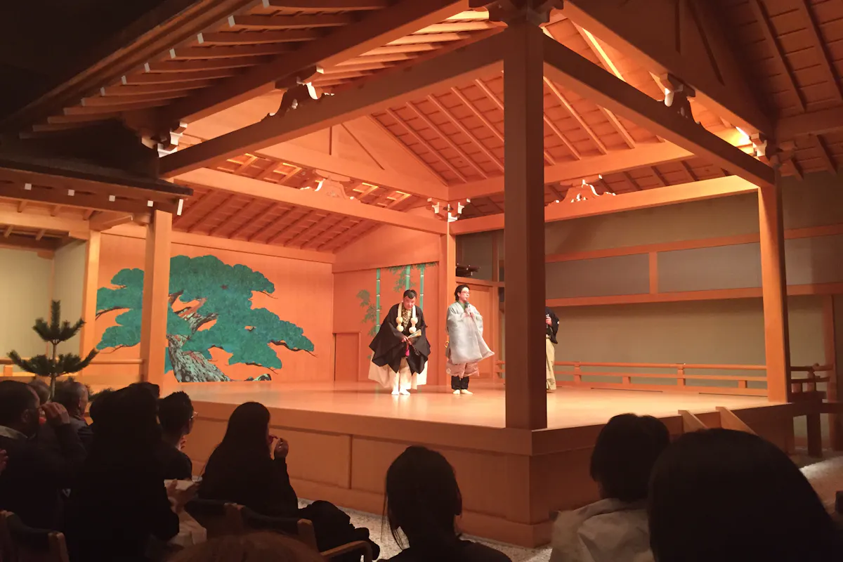 Noh Theatre A traditional Japanese Noh theater performance is taking place on a wooden stage with actors dressed in traditional attire. The backdrop features a green painting of a tree. An audience watches attentively from their seats.