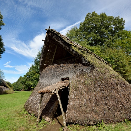 Jomon-Period A traditional thatched-roof hut with a triangular shape stands in a grassy area surrounded by trees, under a blue sky with scattered clouds. Another similar hut is visible in the background.