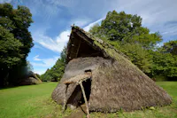 A traditional thatched-roof hut with a triangular shape stands in a grassy area surrounded by trees, under a blue sky with scattered clouds. Another similar hut is visible in the background.
