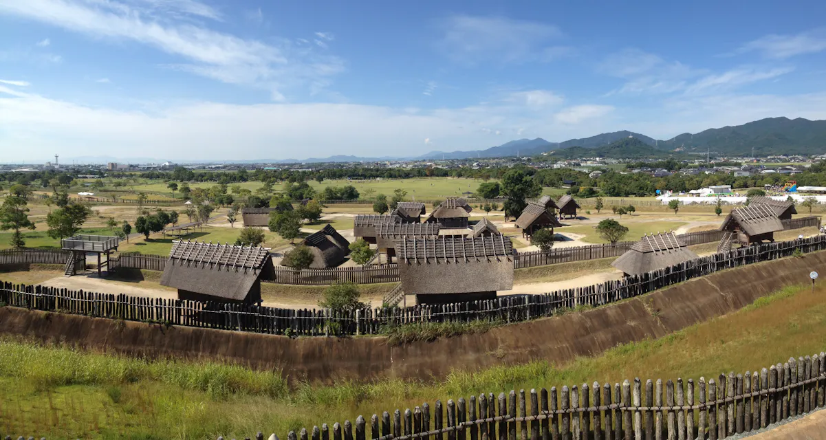Panoramic view of Yoshinogari Historical Park in Japan, showcasing a reconstruction of an ancient Yayoi period village with thatched-roof buildings and wooden fences. The scene is set against a backdrop of rolling hills and a clear blue sky.