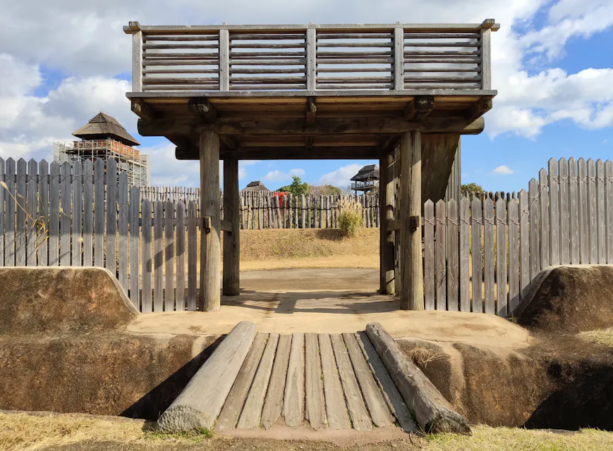 A wooden gate with a roof stands at the entrance of a historic fort, featuring a small bridge over a dug-out trench. The fort is surrounded by a high wooden fence, and watchtowers are visible in the background under a partly cloudy sky.