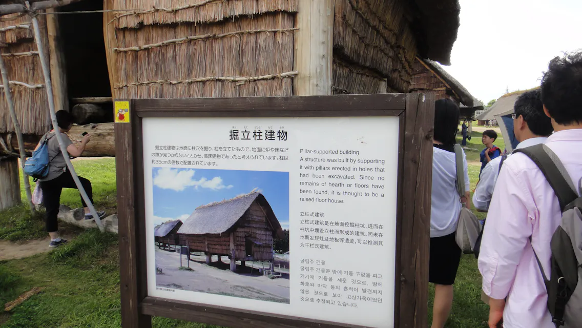 A group of people stands near a sign with text and an image in front of a traditional thatched building. The sign features both Japanese and English text, describing the pillar-supported building, and includes a photo of similar structures.