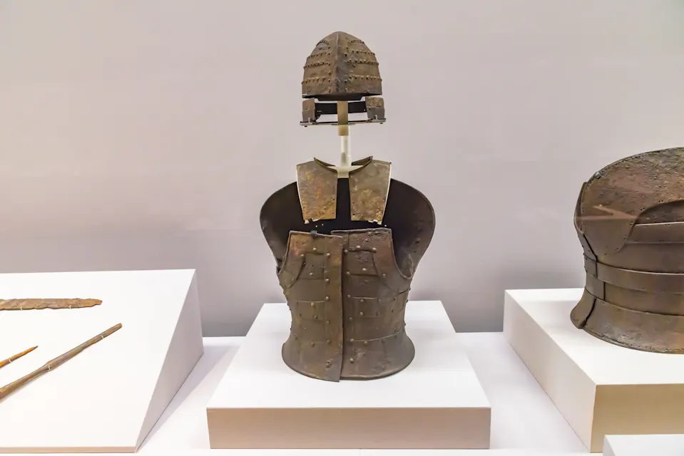 Ancient Japanese helmet and armor exhibit displayed on a white platform. The central piece includes an iron helmet and segmented chest armor, with additional armor pieces and weapons displayed on the left.