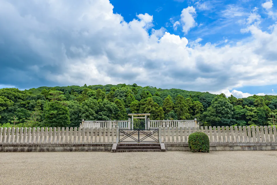 A scenic view of a traditional Japanese torii gate on a hill, surrounded by lush green trees and framed by a white fence in the foreground. Above, a partly cloudy sky contrasts with patches of vibrant blue.