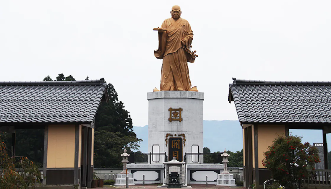 A bronze statue of a man on a pedestal in a park, representing Nichiren, a Buddhist priest in Niigata City, Japan. A large golden statue of a robed figure stands on a tall pedestal between two traditional-style gates with tiled roofs. The outdoor scene is set against a backdrop of trees and mountains under a cloudy sky.