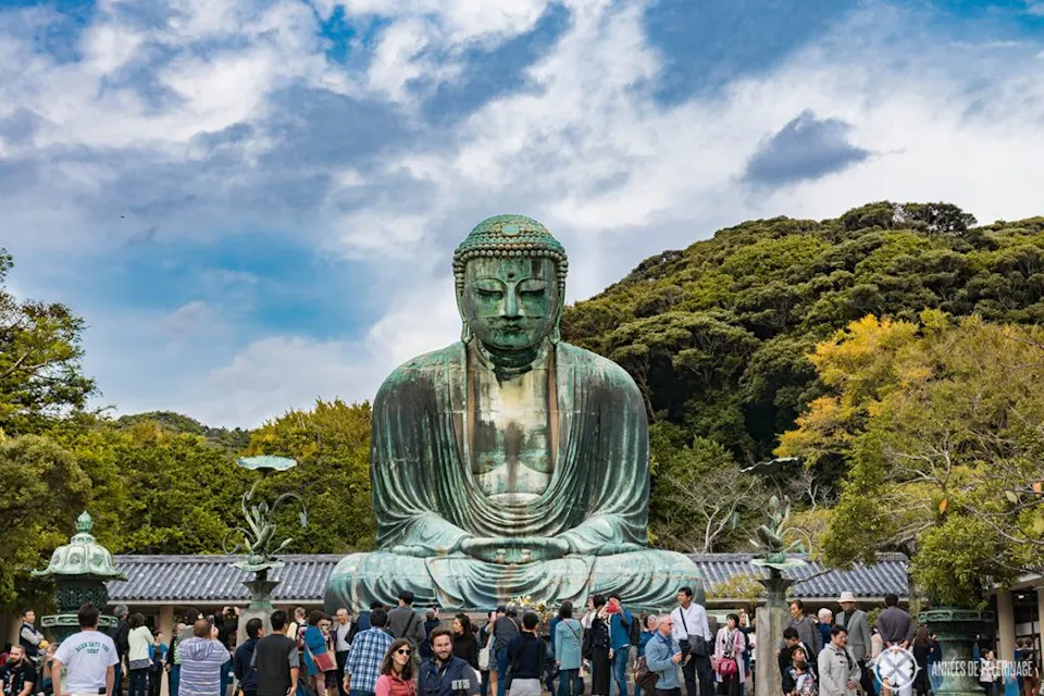 A majestic Buddha statue stands before a crowd with a cloudy sky at back, symbolizing serenity and spirituality at the Kotokuin Temple in Kamakura City, Japan. A large bronze statue of the Great Buddha (Daibutsu) sits outdoors against a backdrop of trees and a cloudy sky. Many people are gathered around the statue, taking photos and sightseeing. The statue is seated in a meditative pose.