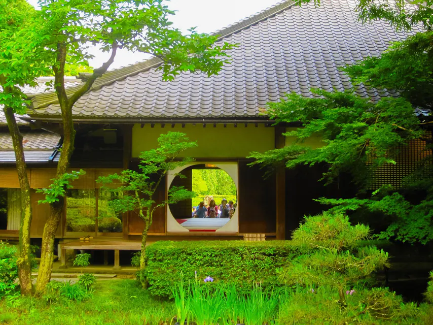 Meigetsu-in Temple in Kamakura, Japan, surrounded by greenery, features a building with a large round passage. A traditional Japanese building with a tiled roof is surrounded by lush greenery. A circular window frames a group of visitors, creating a serene and picturesque scene. In the foreground, there are trees and a small garden with various plants.