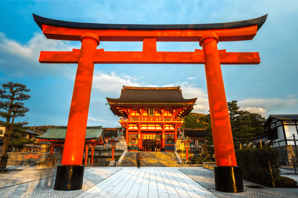 A majestic symbol of Japan's rich heritage, a vibrant red Torii gate stands proudly before a serene Shinto Shrine A vibrant red torii gate stands prominently in front of Fushimi Inari Shrine in Kyoto, Japan, framing the main shrine building. The sky is partly cloudy, and the sun casts warm light on the structures. Lush greenery surrounds the shrine, enhancing its serene atmosphere.