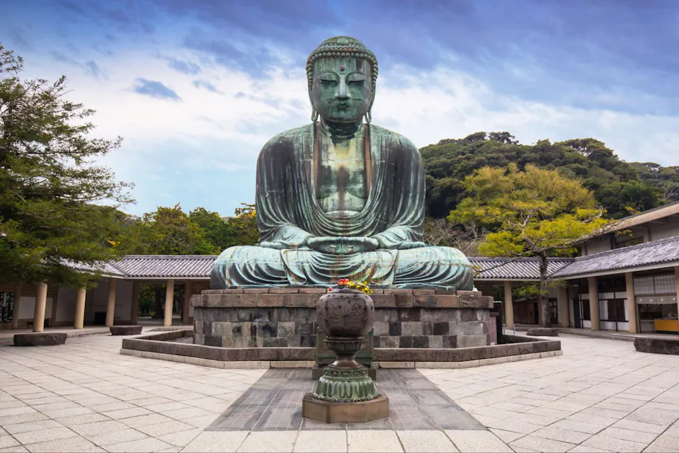 A majestic Buddha statue stands before a crowd with a cloudy sky at back, symbolizing serenity and spirituality at the Kotokuin Temple in Kamakura City, Japan. A large bronze statue of Buddha sits serenely in an outdoor courtyard, surrounded by traditional Japanese buildings. The sky is partly cloudy, and trees with green and yellow foliage frame the statue, adding a touch of nature to the tranquil scene.