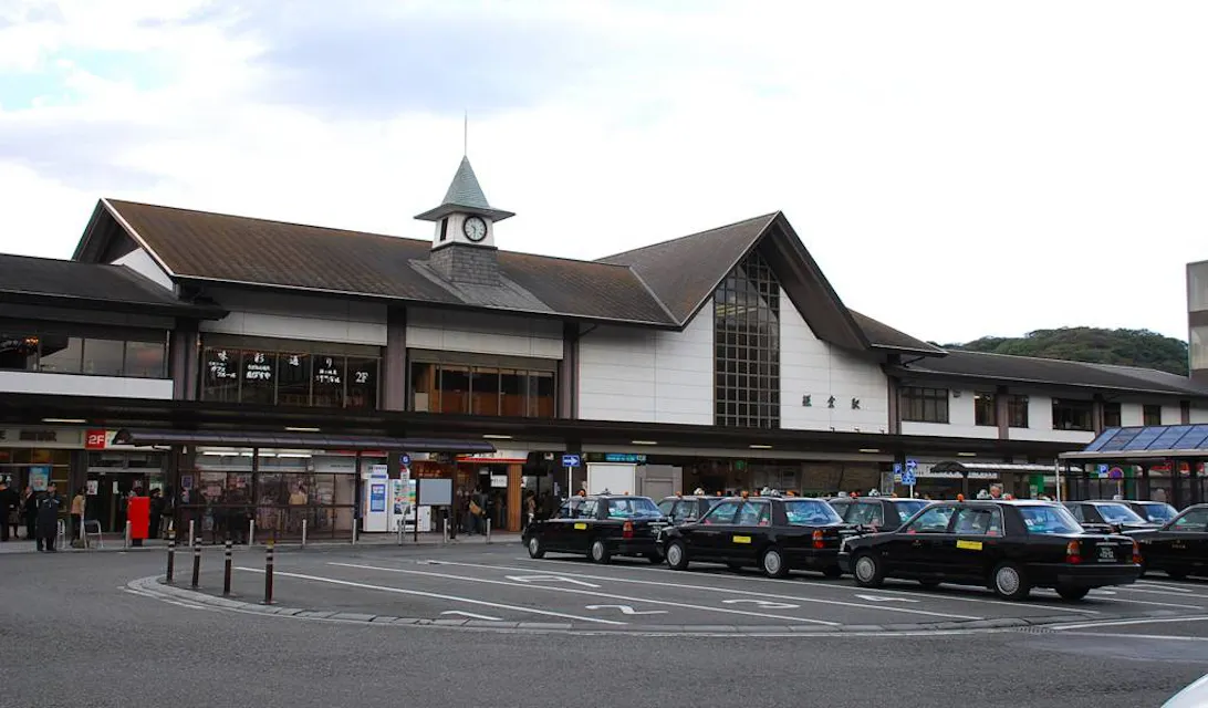 A little clock tower adorns the grand Kamakura Station Area, a symbol of elegance in Japan's Kamakura City. A train station building with a clock tower and gabled roof design. Several black taxis are lined up in front of the station, and people are seen near the entrance. The sky is overcast.