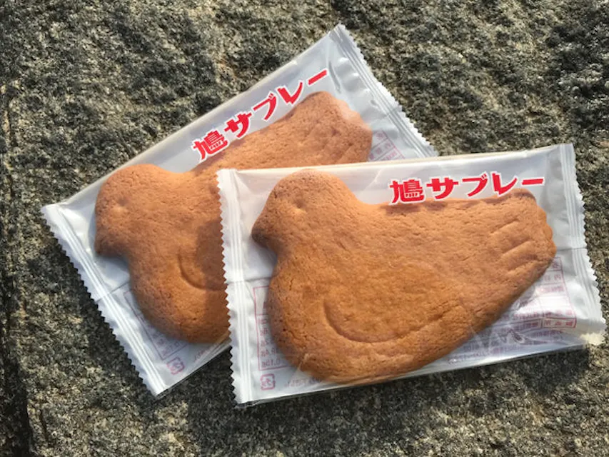 Two delectable cookies adorned with a charming bird design, representing the iconic Kamakura pigeon scone, a beloved local treat. Two bird-shaped cookies wrapped in clear plastic packaging with red Japanese text on top are placed on a stone surface.