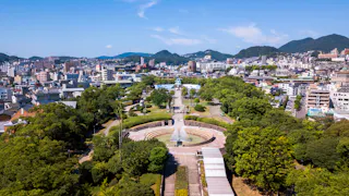 Aerial view of a city park with walking paths, a circular fountain, abundant green trees, and surrounding urban buildings, with distant hills under a clear blue sky.